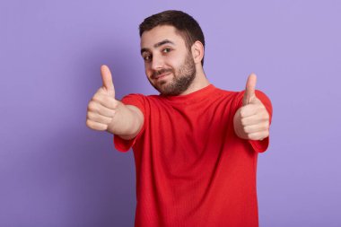 Leylak arkaplanının üzerinde izole edilmiş, sıradan kırmızı tişört giyen genç bir adamın yatay görüntüsü mutlu bir yüz ifadesiyle gülümsüyor, kameraya baş parmağını gösteriyor. Halk konsepti.