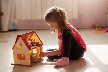 Indoor shot of liitle charming girl sits on floor with her doll house, wearing casual clothing, has ponytail. Self isolation during coronavirus pandeic. Children spends time at home duing quarantine. clipart