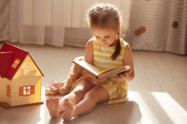 Oyuncak bebek evinin yanında yerde otururken oyuncağı için kitap okuyan sevimli küçük kız. Karantina sırasında evde hoş bir çocuk, koronavirüs salgını sırasında kendini izole ediyor. Covid 19 konept.