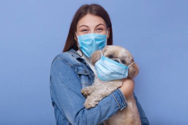 Kot ceket giyen, maske takan ve mavi stüdyo arka planında izole edilmiş poz veren köpek taşıyan bir kadının yatay görüntüsü. Coronavirüs salgını, qaurantine, çevre kavramı.