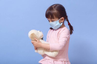 Günlük giysiler ve tıbbi maske takan küçük bir kızın portresini yakından çek. Yavru köpek oyuncağını elinde tut ve ona bak. Mavi stüdyo arka planında saçı örgülü bir kadın çocuğu. Coronavirüs.