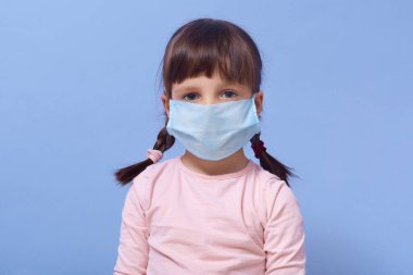 Roseshirt giyen ve yüzüne tek kullanımlık maske takan küçük bir kız resmi. Mavi arka planda izole bir şekilde poz veriyor, kameraya bakıyor, üzgün bir bakış atıyor. Coronavirus salgını koruması, covid 19 konsepti..