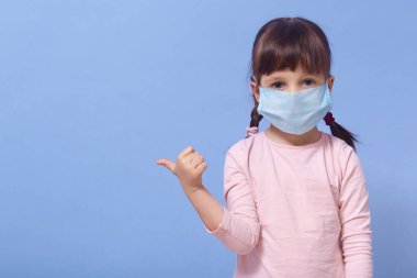 Küçük kızın tıbbi koruyucu maske ve gül tişörtü giyip stüdyoda poz verirken başparmağıyla işaret ettiği yatay bir görüntü. Coronavirus, covid 19, karantina konsepti. Reklam alanını kopyala.
