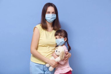 Koronavirüs konsepti, karantina, covid 19. Anne ve kızı tıbbi maskeler takıyor, birbirlerine sarılıyorlar ve doğrudan kameraya bakıyorlar. Küçük kız en sevdiği oyuncağını mavi duvarın üzerinde tutuyor..