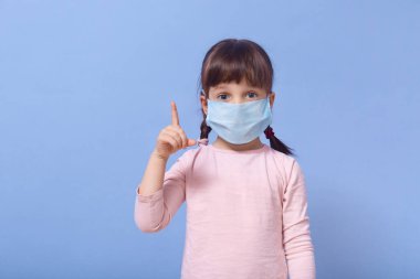 Açık gözlü, sıradan bir gül gömleği ve koruyucu bir maske takmış işaret parmağıyla işaret ederek dikkat çeken küçük bir kız. Koronavirüse karşı koruma, covid 19 konsepti.