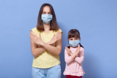 Coronavirus salgını sırasında tıbbi koruyucu maskeler takan anne ve küçük kız görüntüsü mavi arka planda çapraz kollarla durdurma hareketini gösteriyor..