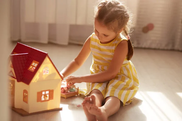 Little girl playing with doll house while siiting on floor in bedroom. Role game for young children. Female child putting to bed her toy doll, charming little lady wears yellow dress. Children concept