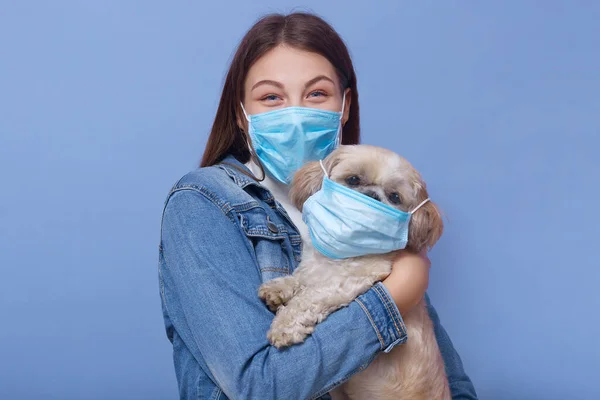 Horizontal shot of woman wearing denim jacket, face mask and hoding dog with flumask too, posing isolated over blue studio background. Coronavirus pandemic, qaurantine, environmental concept.