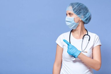 Kafkas doktor kadının resmi mavi stüdyo arka planında izole edilmiş, şapka, eldiven ve flüt maske takıyor, işaret parmağıyla işaret ediyor, yan bakıyor. Reklam veya tanıtım için alanı kopyala.