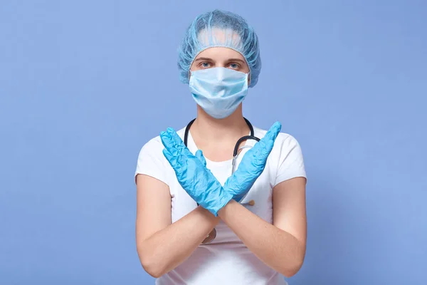 Picture of woman wearing medical protection mask, medical hat and rubber gloves, crossing hands to stop virus, epidemy, world pandemia, posing isolated over blue studio background. Health care concept