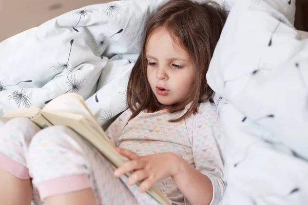 Profile of hard working curious smart girl learning to read, looking at text attentively, reading in voice, holding book, having fair hair, staying in bed, being on weekends at home. Study concept.