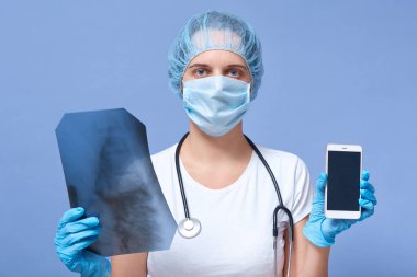 Kapalı alanda, elinde röntgen taraması olan ve teşhis merkeziyle telefonda konuşmaya hazır olan, mavi stüdyo arka planında izole edilmiş poz veren, kameraya bakan kadın doktorun steteskopu var. Boşluğu kopyala.