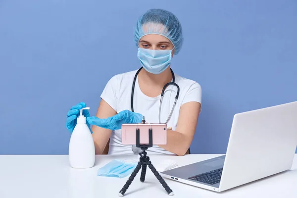 Close up portrait of medical doctor works online from home, helping people in days of panic and pandemic, shows in her vlog how to use sanitaizer, sitting at white desk in front of tripod with phone.