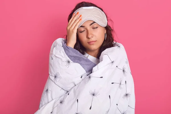 Horizontal shot of young girl with fresh skin, poses with closed eyes, has blindfold on her forehead, keeping hand on her forehead, looks sleepy, wrapped white blanket against rosy studio wall.