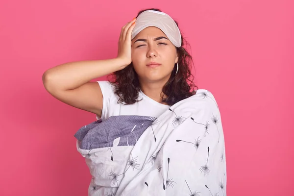 Close up portrait of dark haired lady with bad look, female has terrible headache,keeping hand on her temple, standing wrapped in white blanket, woman looks sick, isolated over rosy studio background.