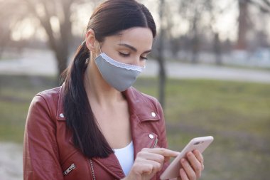 Siyah saçlı, at kuyruklu, avluda dikilmiş, cep telefonunu elinde tutan arama numarası, konsantre olmuş, koruyucu maske takan bir kadın. Corona virüsü salgını konsepti.