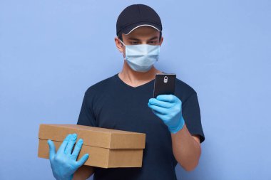 Akıllı telefonu elinde olan teslimatçının yatay görüntüsü. Yazılı teslimat adresi ya da telefon görüşmesi. Elinde kutuyla Corona virüsünün yayılmasını önlemek için koruyucu giysiler giyiyor. .