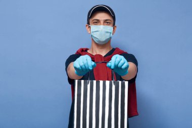 Closeup portrait of courier handing stripped shopping bag with online purchase, delivery man waring mask and gloves to prevent spreading dangerous corona virus all over world. Covid 19 concept. clipart
