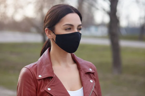 Outdoor shot of beautiful brunette woman wearing leather jacket and black protective mask, looking in distance, protecting herself from dangerous corona virus, walking in open air. Covid 19 concept.
