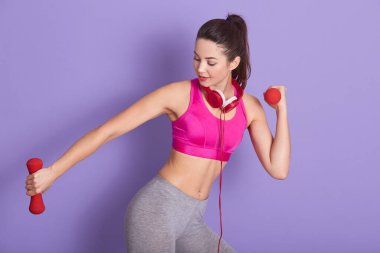 Sportif görünümlü spor eğitmeninin fiziksel egzersiz yaptığı, kırmızı halterler tuttuğu, kulaklık taktığı, pembe bluz giydiği ve gri tayt giydiği bir resim. İnsanlar ve spor konsepti.
