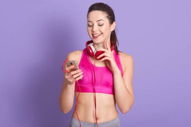 Güzel, neşeli bir bayanın kapalı bir portresi samimi bir şekilde gülümsüyor, hoş bir yüz ifadesi takınıyor, cihaz ekranına bakıyor, akıllı telefon tutuyor, spor giysiler giyiyor. Spor antrenörü konsepti.