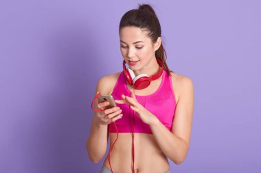 Yakından bakınca, atkuyruklu, pembe spor sutyen giyen, spor yapmak için müziği seçen, cihazın ekranına bakan, eflatun rengi stüdyo arka planında izole edilmiş poz veren, sevimli, koyu renk saçlı bir kadının portresi..