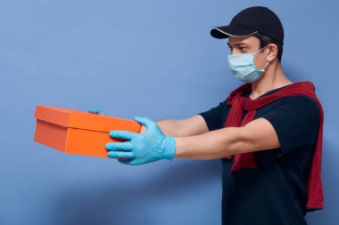 Kuryenin yüz maskesi ve lateks eldiven taktığı, müşterisine karton kutu verdiği, mavi stüdyo arka planına izole edilmiş poz verdiği, teslimatçı adamın koronavirüs karantinası sırasında çalıştığı kapalı çekim..