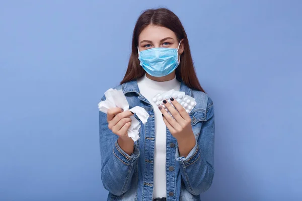 室内拍摄的生病女士身穿休闲装 戴着防护面具 在蓝色的工作室墙壁上摆出隔离的姿势 手里拿着药丸 直接看着相机 Corona Virus Health Care Concept — 图库照片