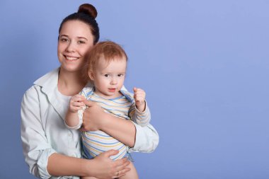 Mutlu gülümseyen annenin küçük oğlunu ellerinin arasında tutarken, çizgili mayo giymiş bir bebek yumruklarını gösterirken, mavi arka planda izole bir şekilde poz veriyor. Reklam veya tanıtım için boşluk kopyala