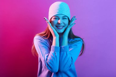 Gündelik kazak ve şapka giyen mutlu bir gencin yatay görüntüsü, iyi bir ruh hali içinde, avuçlarını yanaklarında tutuyor, kameraya gülümsüyor, izole pembe neon stüdyo arkaplanı pozu veriyor.