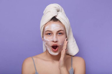 Kolsuz tişört giyen, beyaz havluya sarılmış, ağzı kapalı, dudaklarını yuvarlak tutan, jöle ile temizleme prosedürleri yapan, mavi duvarda izole edilmiş bir şekilde duran sevimli kızın stüdyo fotoğrafı..