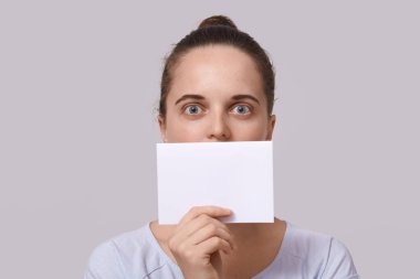 Stüdyoda, saçı topuzlu, dudaklarının yanında boş bir kart tutan, büyük gözlerle poz veren, dudağını, yazıtlar için fotokopi alanı olan bir kağıt parçasıyla kaplayan, stüdyo arka planında izole edilmiş bir kadının fotoğrafı..