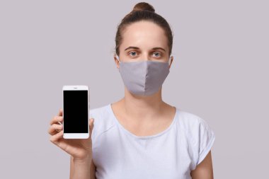 Ciddi yüz ifadesi olan, elinde boş ekran olan akıllı bir telefonu tutan, kameraya bakan, yüz maskesi takan, beyaz arka planda izole bir şekilde poz veren bir kadının stüdyo fotoğrafı. Corona virüsü konsepti.