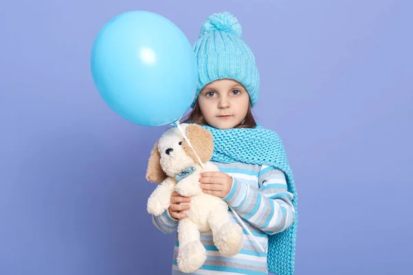 Closeup portrait of charming little female child wearing stripped shirt, turquoise scarf and hat, posing isolated over lilac studio background, holding balloon and soft dog toy. Childhood concept.