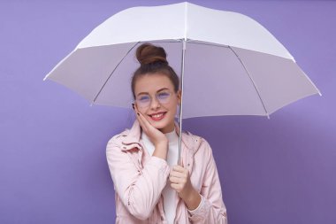 Neşeli, neşeli, çekici genç bir kadının portresi. Elini yanağına koyuyor, şemsiye tutuyor, hoş bir yüz ifadesi takınıyor, içtenlikle gülümsüyor, pembe ve beyaz elbiseler giyiyor. Ruh hali konsepti.