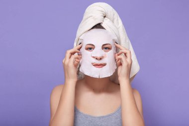Yüzüne maske takan, cildini nemlendiren, kendini önemseyen, kafasında havlu olan, stüdyodaki leylak arkaplanının üzerinde tek başına duran güzel bir kadın resmi. Kadınlar ve güzellik konsepti.