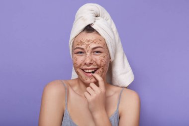 Kafasında havlu olan mutlu genç bir kadının yatay çekimi stüdyodaki leylak arkaplanının üzerinde izole bir şekilde duruyor, parmaklarını ağıza veriyor, doğrudan kameraya bakıyor. Duygular konsepti.