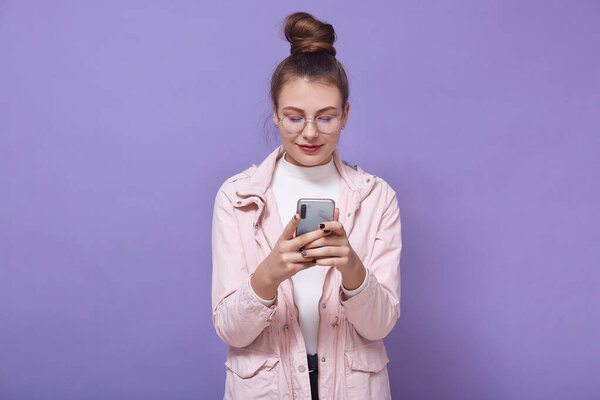 Portrait of pleasant magnetic good looking attractive teenager holding smartphone in hands, spending time alone, having addiction, wearing leather jacket, sweater and eyeglasses. Youth concept.