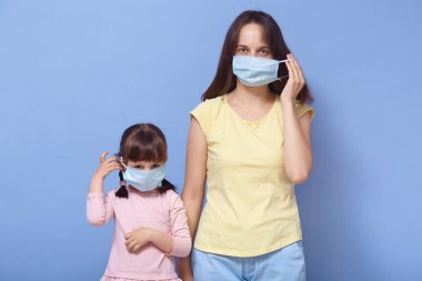Kızıyla birlikte stüdyoda mavi arka planda duran, tıbbi maskeler takan, doğrudan kameraya bakan, evde karantinada olan genç bir kadının yatay görüntüsü..