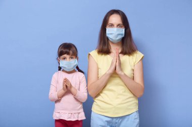Ciddi odaklanmış genç bir kadının portresi. Küçük kızı stüdyoda mavi arka planda tek başına duruyor. El ele tutuşuyor, dua ediyor, tıbbi maskeler takıyor. Evde kal konsepti.