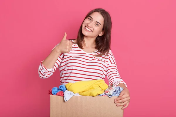 Horizontal shot of happy brunette woman showing thumb up and looking directly art camera, wearing casual attire, holding box with clothes for secondary using, female volunteer helping poor people.