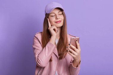 Stüdyo çekimlerinde hoş bir bayan soluk gül ceketi ve şapka giyer, akıllı telefonu elinde tutar ve ekranına bakar, işaret parmağını yanağında tutar, olumlu duygular ifade eder, gülümseyerek poz verir..