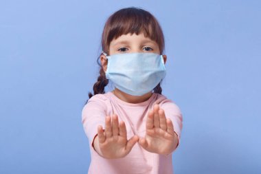 Yakından bakınca, günlük gömlek ve tıbbi maske takan sevimli bir çocuğun portresi, iki avucunda da dur hareketi yapan, kameraya bakan, iki örgülü, mavi stüdyo arka planında izole edilmiş poz veren kız çocuğu..