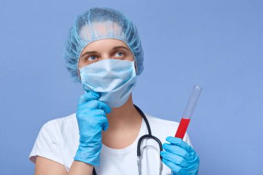 Kapalı alanda, düşünceli bir doktorun muayene ederken, cam tüpte kan örneği tutarken, yana bakarken, düşünceli bir yüz ifadesi takınırken, elini çenenin yakınında tutarken. Tıp, sağlık hizmeti kavramı.