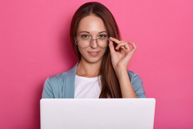 Hoş görünümlü, gülümseyen bir kızın kapalı çekimleri, gözlüklerinin çerçevesinde elini tutan bir kadın, kameraya bakıyor, pembe stüdyo duvarına karşı poz veriyor. Taşınabilir bilgisayarda çalışan serbest çalışan.