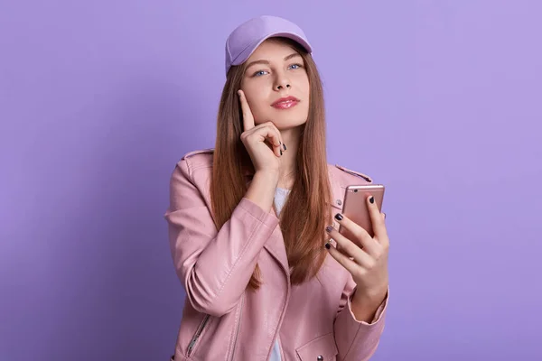 Studio shot of charming lady looking directly at camera while holding smart phone in hands and keeping finger on cheek, student girl posing isolated over lilac studio background. People concept.