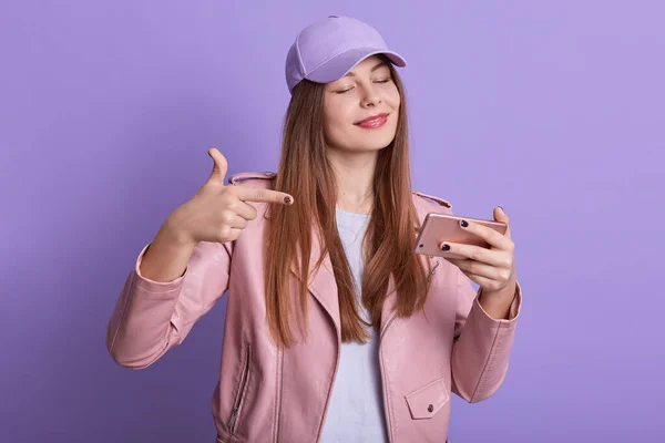Horizontal shot of attractive female wearing casual attire and cap, standing against lilac background and pointing at device screen while has video call with friend, lady with pleasant appearance.