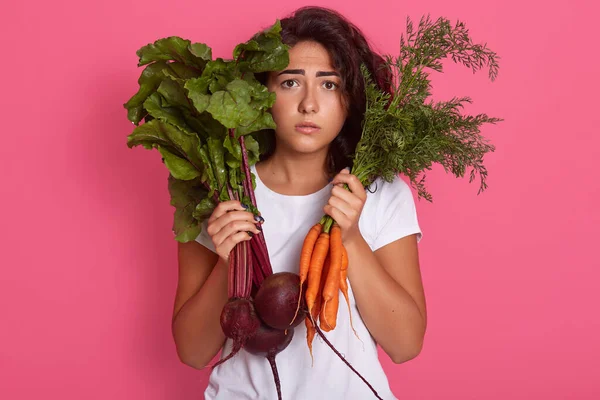 Closeup portrait of upset woman holding in hands carrots and beetroots, female wearing white casual t shirt, girl with upset facial expression, has negative emotions, stands against pink studio wall.