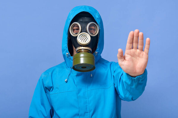 Studio shot of man dresses chemical protective suit and respirator posing and showing stop gesture with his hand against blue wall. Passage is forbidden. Air pollution, danger of infection concept.