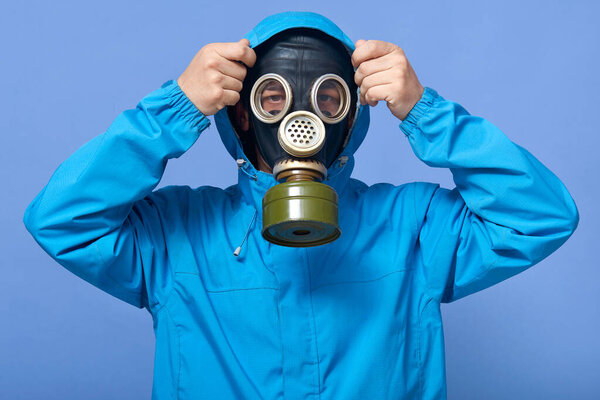 Closeup portrait of man putting on hood, male wearing respirator on his face and uniform, scientist fights with pollution of environment, guy posing against blue studio wall. Ecology concept.